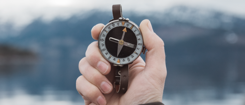 Eine Hand hält im Freien einen Zirkel, im Hintergrund sind verschwommen Wasser und schneebedeckte Berge zu sehen.