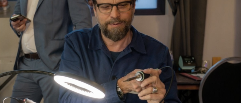 Ein Mann mit Brille und Bart hält an einer beleuchteten Werkbank einen Lötkolben in der Hand, während im Hintergrund eine andere Person im Anzug steht und telefoniert.